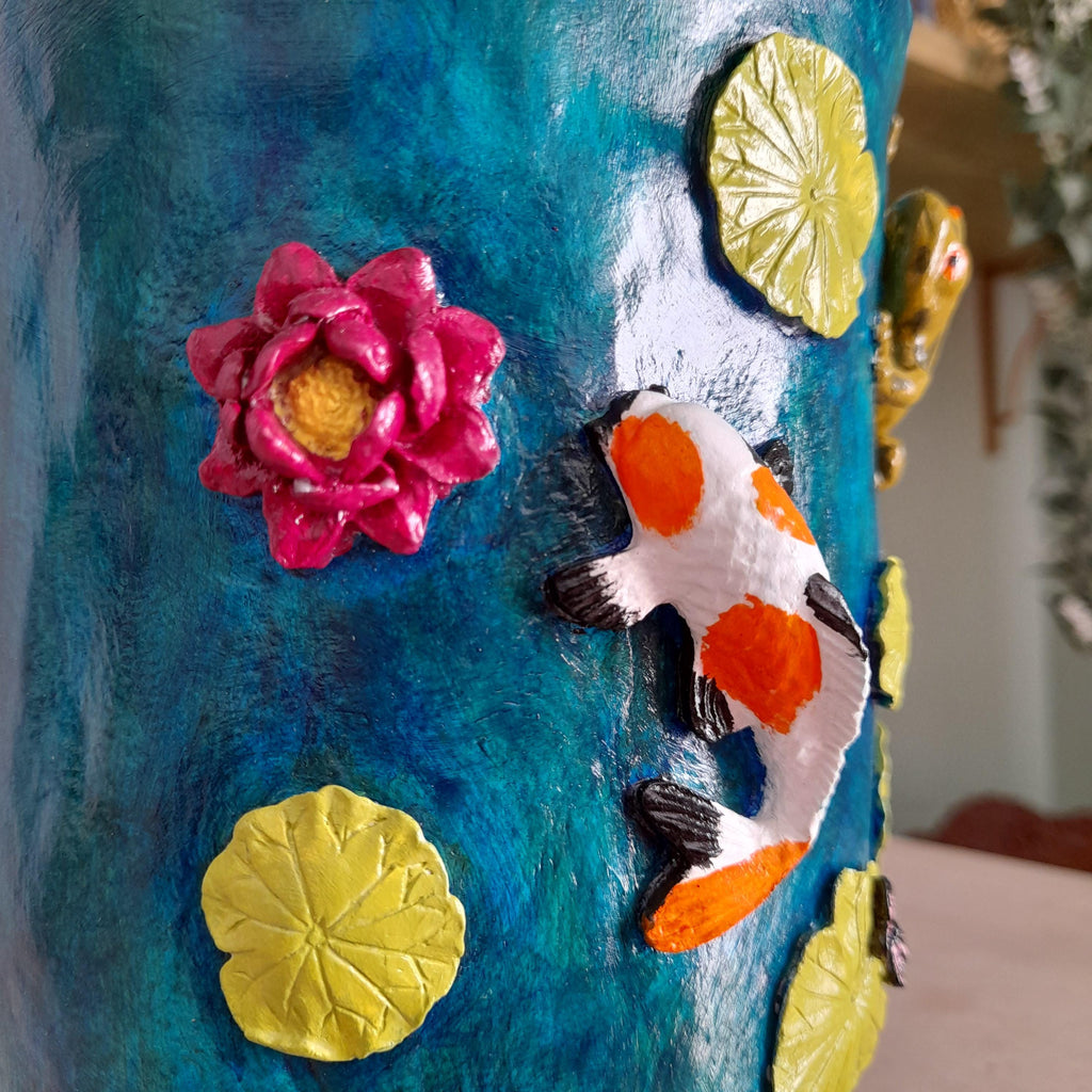 A close-up of a decorative cylindrical surface featuring a textured blue background with raised 3D elements including a pink lotus flower with a yellow center, yellow lily pads, and a white koi fish with orange and black markings.