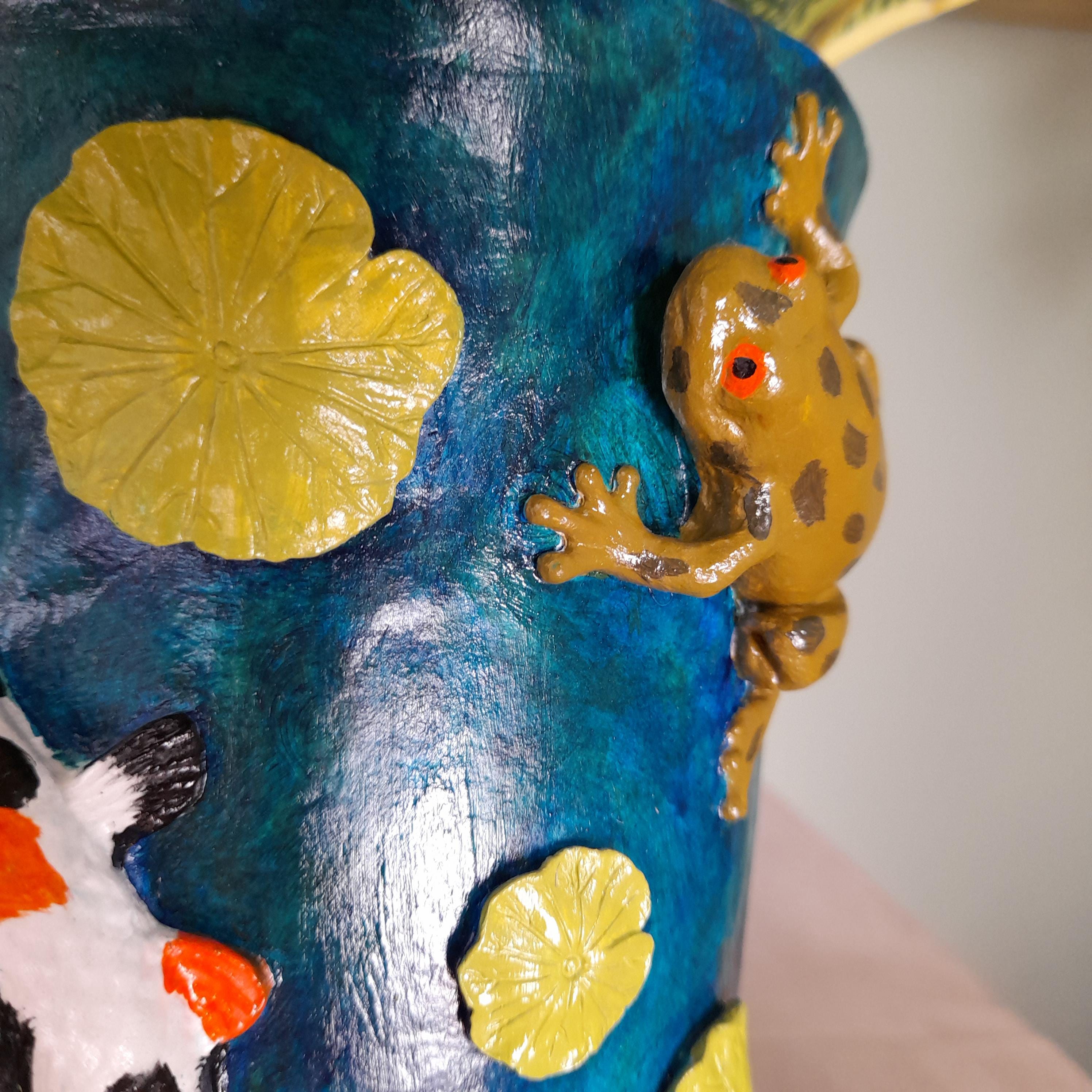 A close-up of a hand-painted decorative pot featuring a blue watercolor-style background with yellow lily pads, a brown frog climbing up the side, and part of a koi fish illustration.