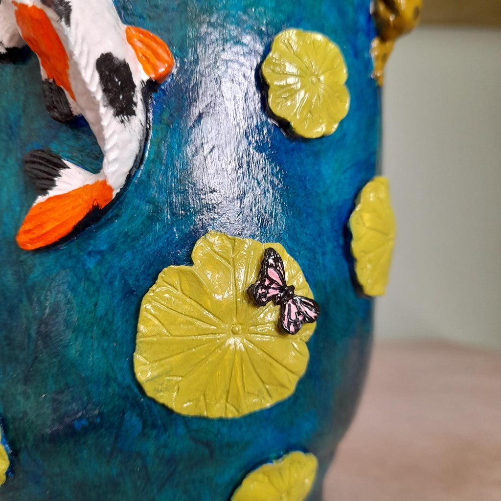 Close-up of a decorative blue ceramic pot or vase with yellow lily pads, a koi fish, and a butterfly ornament.