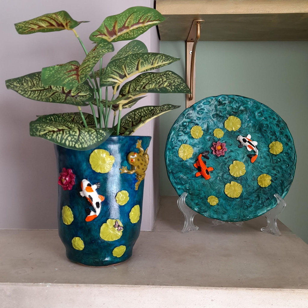 A decorative teal ceramic planter with yellow lily pads and koi fish, holding a green leafy plant, displayed next to a matching round decorative plate on clear stands.