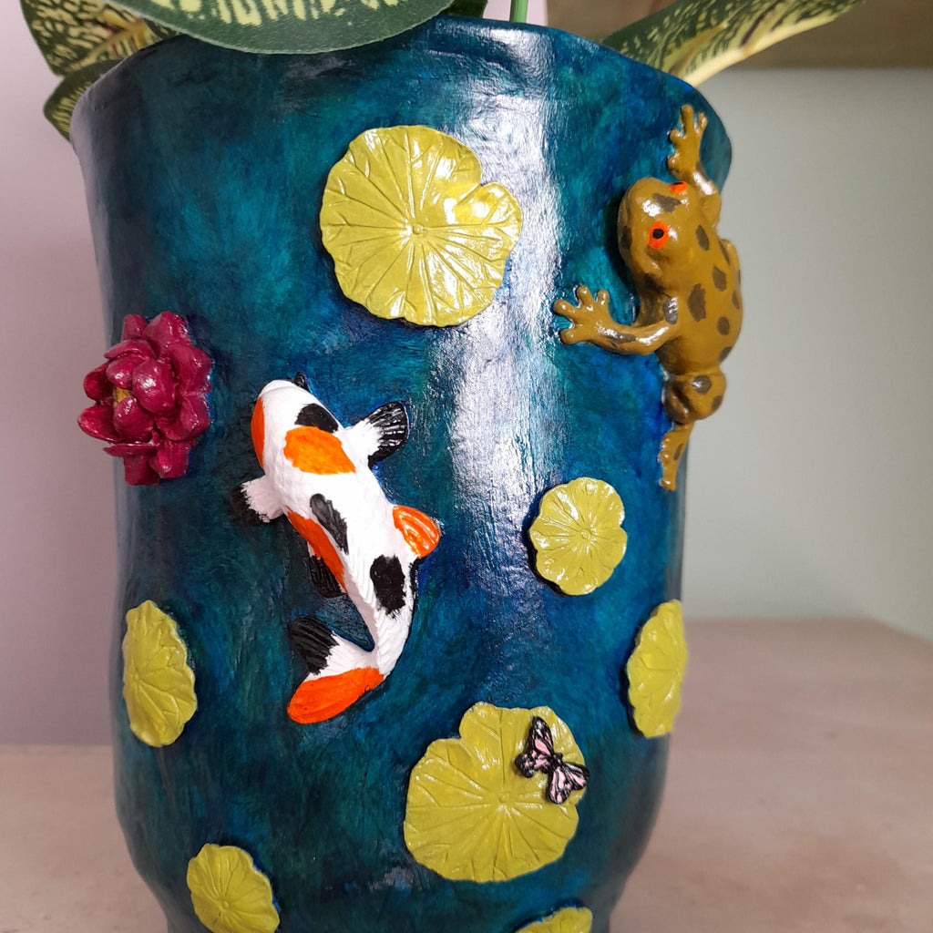 A decorative ceramic planter with a textured dark blue background featuring raised 3D elements of a koi fish, a frog, lily pads, a flower, and a butterfly, shown with green leaves at the top.
